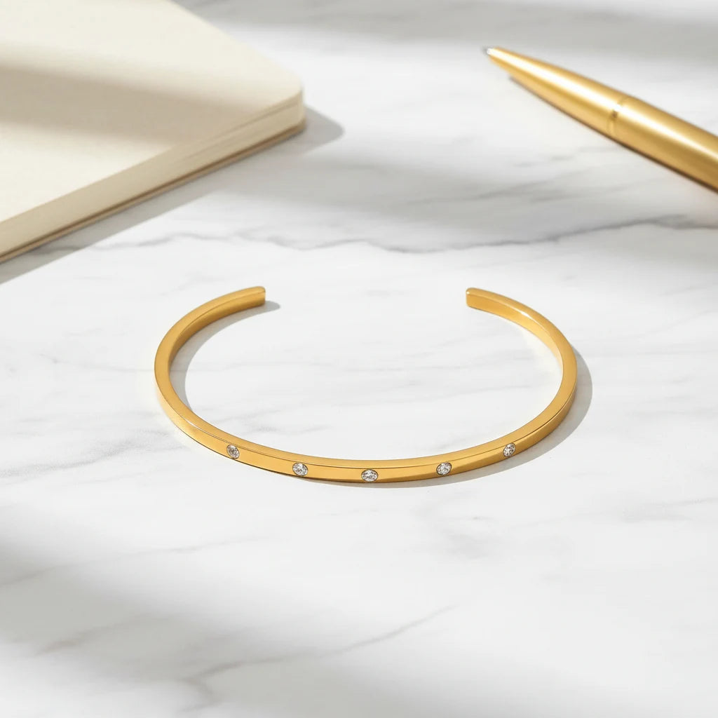 Flat-lay of a thin gold cuff bracelet on white marble next to a cream notebook and gold pen, showcasing its sleek bar shape and sparkling stones for a chic desk look | Oria Jewel