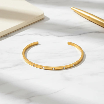 Flat-lay of a thin gold cuff bracelet on white marble next to a cream notebook and gold pen, showcasing its sleek bar shape and sparkling stones for a chic desk look | Oria Jewel
