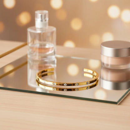 Glam vanity scene with a thin gold cuff bracelet placed on a mirrored tray beside perfume and cosmetics, with warm bokeh lights in the background creating an evening-ready sparkle | Oria Jewel