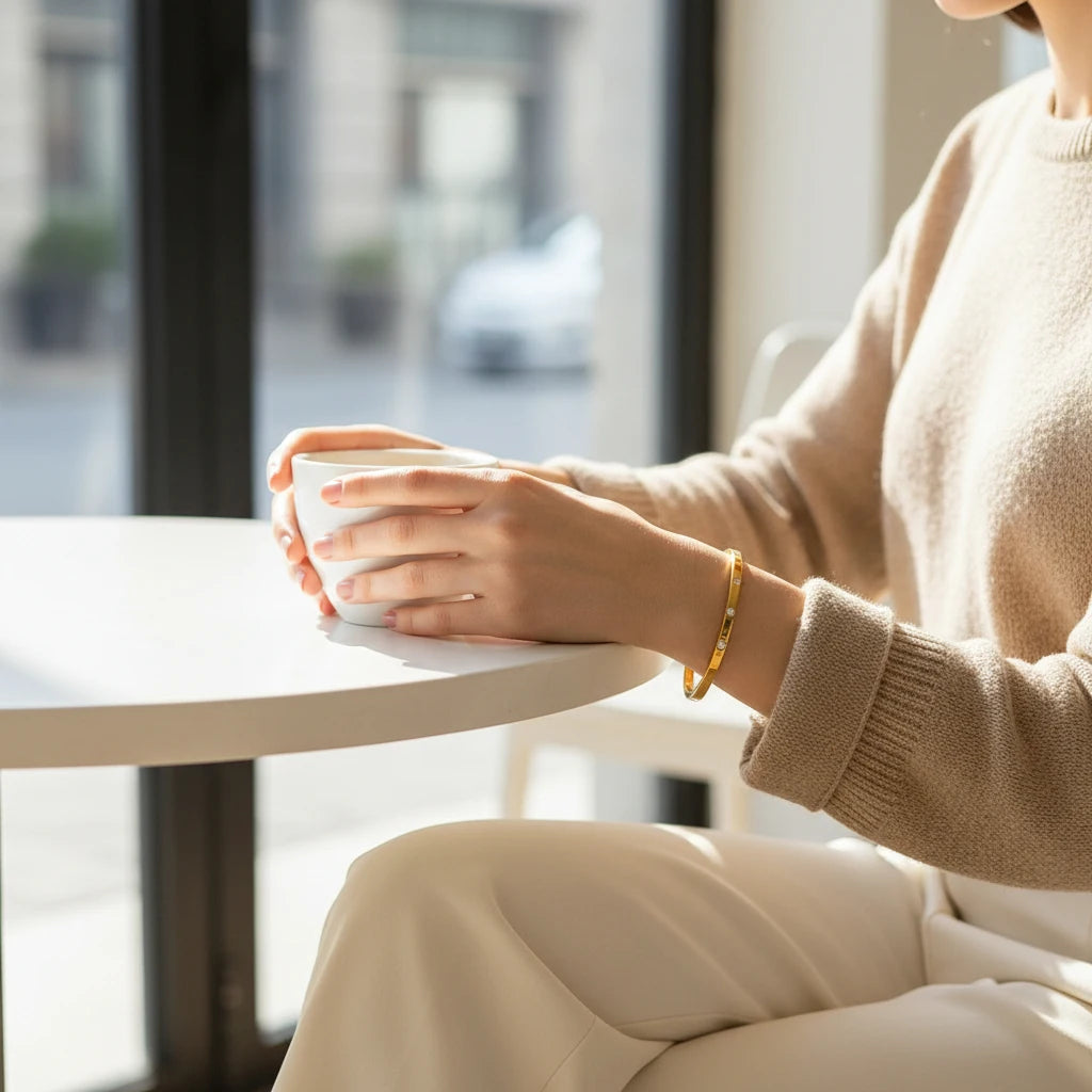 Casual café moment with a woman in a soft beige sweater holding a cup, the thin gold cuff bracelet circling her wrist as a simple, polished everyday accessory | Oria Jewel